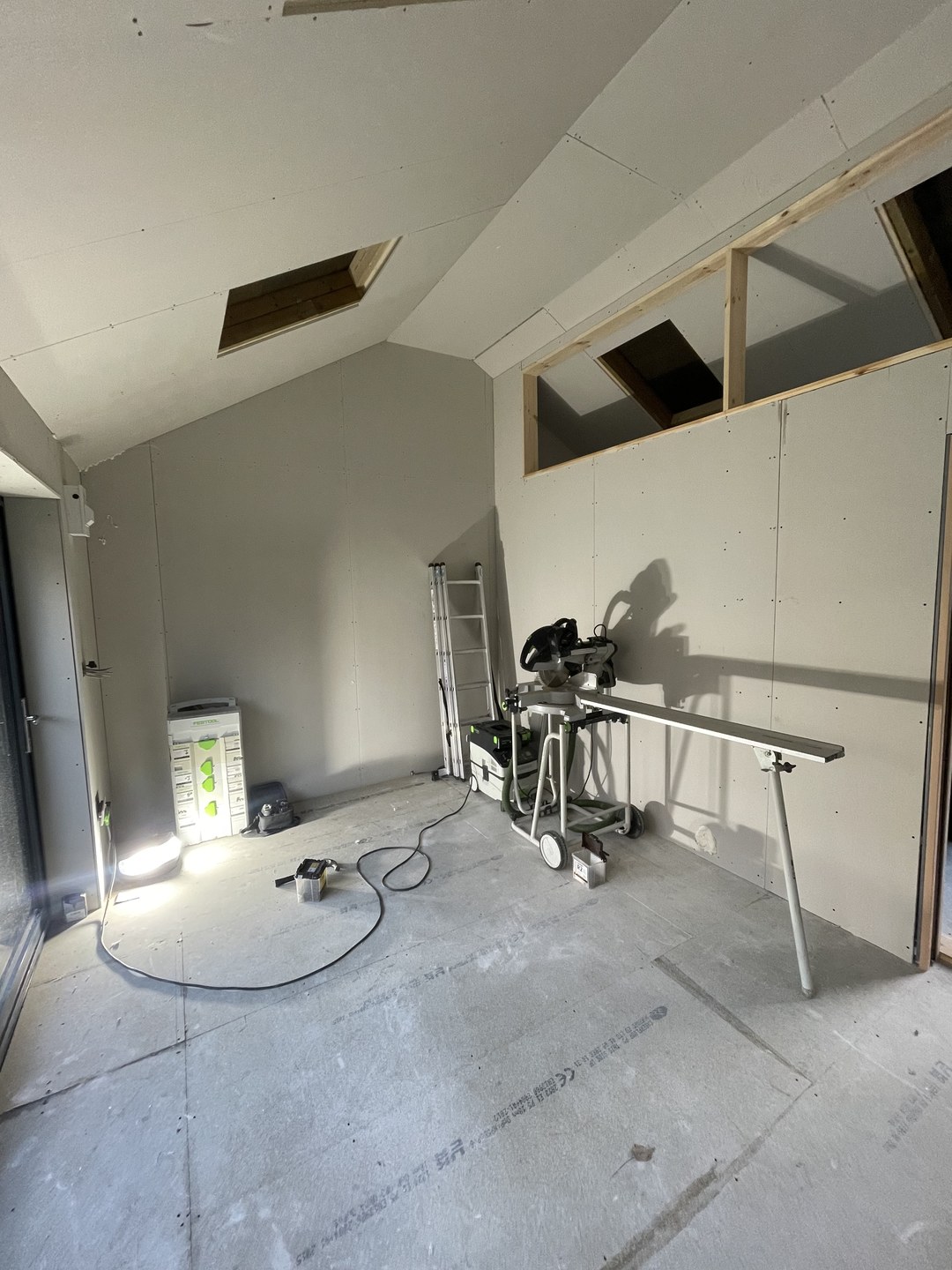 Bare loft room mid-renovation with plasterboard walls, concrete floor, ladder and mitre saw